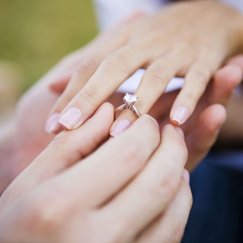 Young man is seen slipping an engagement ring on his girlfriend's ring finger.